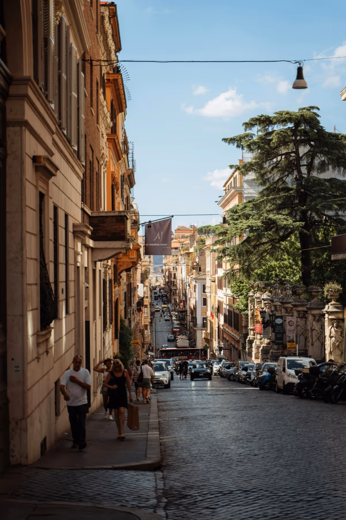 A sunlit cobblestone street in Rome, warm ochre palazzi lining both sides and pedestrians walking downhill.